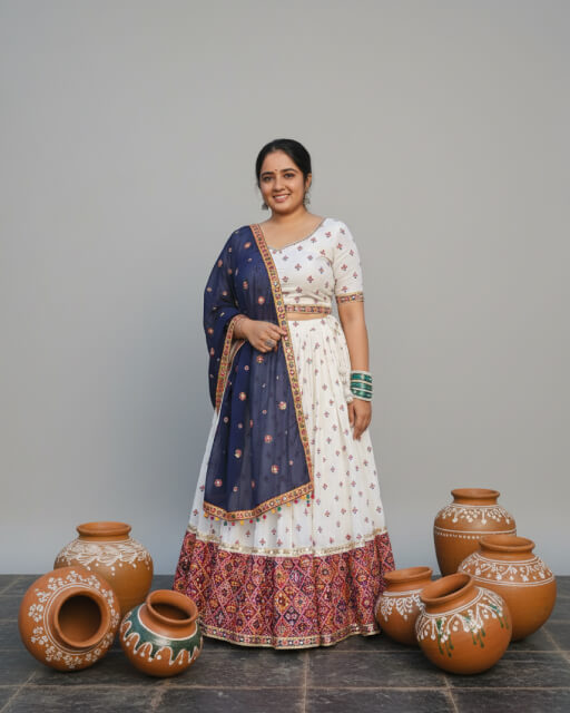 Woman wearing a white chaniya choli with colorful embroidered border and navy blue dupatta, custom-designed by Anokhi by Sneha boutique in Rajkot, surrounded by traditional decorated clay pots.