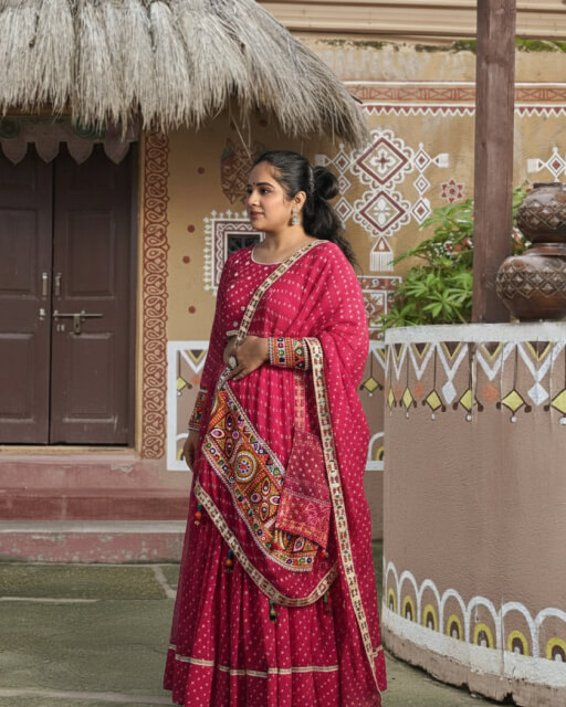 Woman wearing a red Bandhani chaniya choli with Kutchi embroidered dupatta, custom-designed by Anokhi by Sneha boutique in Rajkot, posing against a traditional rural-themed backdrop.